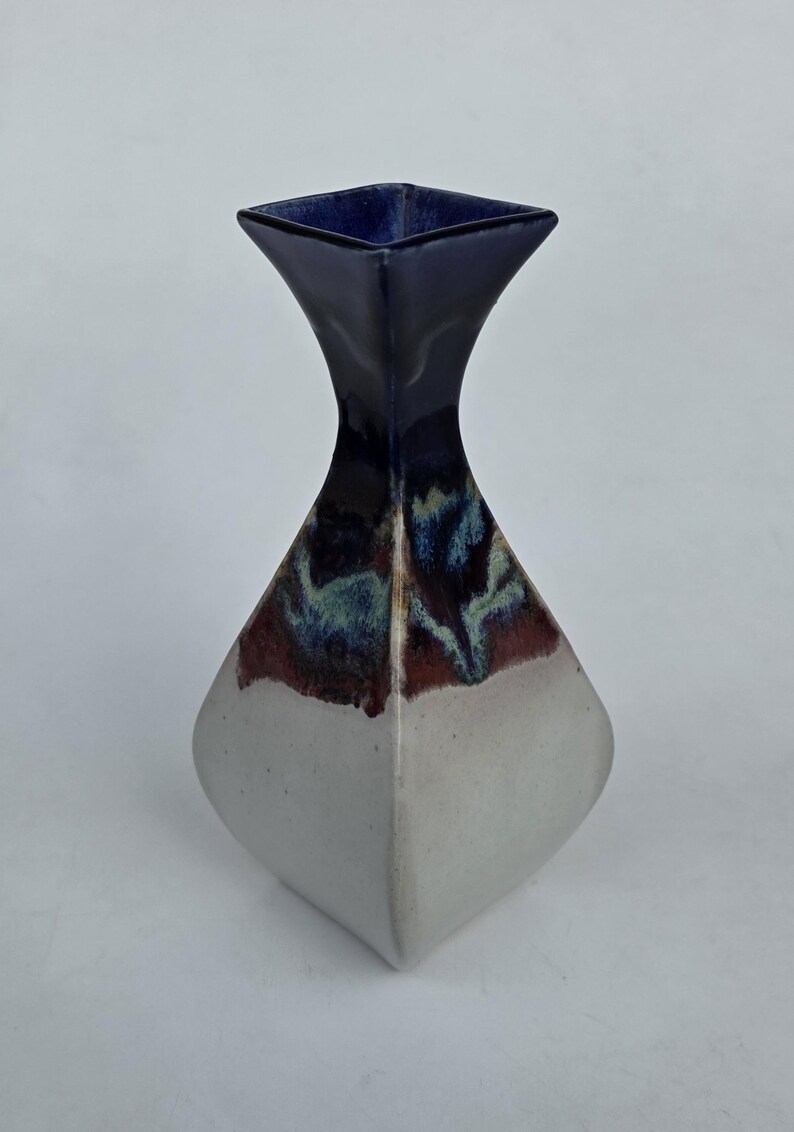 May include: A ceramic vase with a square top and a diamond-shaped body. The top is dark blue, transitioning to a mix of red, blue, and green, and then to a white base. The vase has a modern, geometric design.