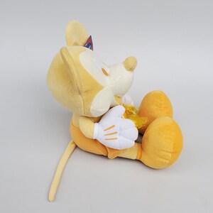 Puede incluir: Un juguete de peluche amarillo de Mickey Mouse sentado con las piernas cruzadas. El juguete tiene una cara blanca y guantes blancos. El juguete tiene una cola amarilla.