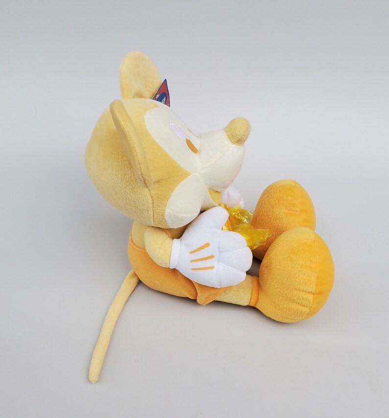 Puede incluir: Un juguete de peluche amarillo de Mickey Mouse sentado con las piernas cruzadas. El juguete tiene una cara blanca y guantes blancos. El juguete tiene una cola amarilla.