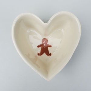 May include: A heart-shaped, off-white ceramic dish with a brown gingerbread man design in the center. The dish has a smooth, glossy finish and is suitable for serving small snacks or as a decorative piece.