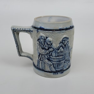 May include: A blue and white ceramic beer stein with a raised design of three men drinking from mugs. The stein has a handle and a rounded base.