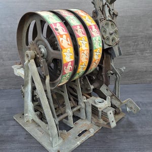 May include: A vintage mechanical slot machine internal mechanism featuring three large rotating metal wheels with colorful printed numbers and symbols, mounted on a weathered gray cast metal frame with exposed gears, levers, and intricate industrial components.