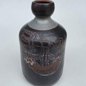 May include: A dark brown ceramic vase with a rounded neck and a wide body. The surface features a textured, etched geometric pattern in a lighter brown tone, with a smooth, matte finish on the neck and a subtle greyish gradient near the rim.