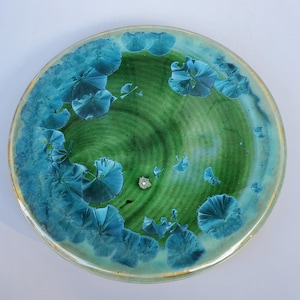 May include: A green and blue ceramic plate with a swirling pattern and a light blue glaze. The plate has a unique, organic design with a speckled finish.