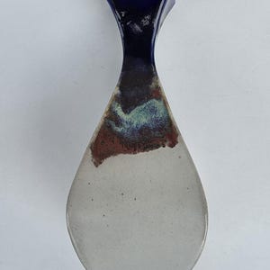 May include: A ceramic vase with a teardrop shape. The top is a deep blue, transitioning to a dark brown and red band, and then to a light gray base. The vase has a unique, modern design.