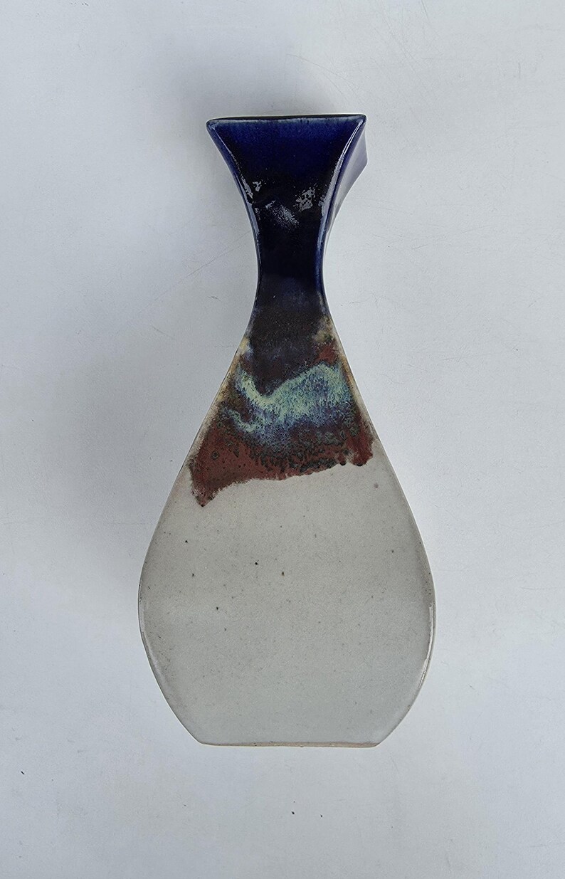 May include: A ceramic vase with a teardrop shape. The top is a deep blue, transitioning to a dark brown and red band, and then to a light gray base. The vase has a unique, modern design.