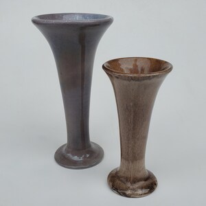 May include: Two ceramic vases with flared rims and rounded bases. The taller vase is a dark purple-brown with a glossy finish. The shorter vase is a lighter brown with a matte finish. Both vases have a classic, elegant shape.