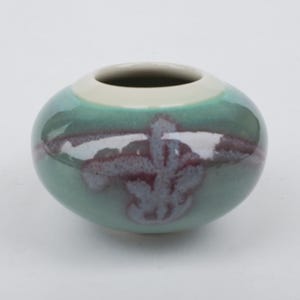 May include: A small, round ceramic vase with a wide opening. The vase is primarily teal with a white rim and a decorative pattern in shades of purple and white. The vase has a glossy finish.