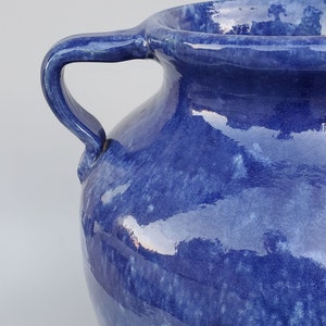 Op de afbeelding: Een blauwe keramische vaas met twee handvatten. De vaas heeft een gevlekt blauw glazuur met een licht iriserende afwerking.
