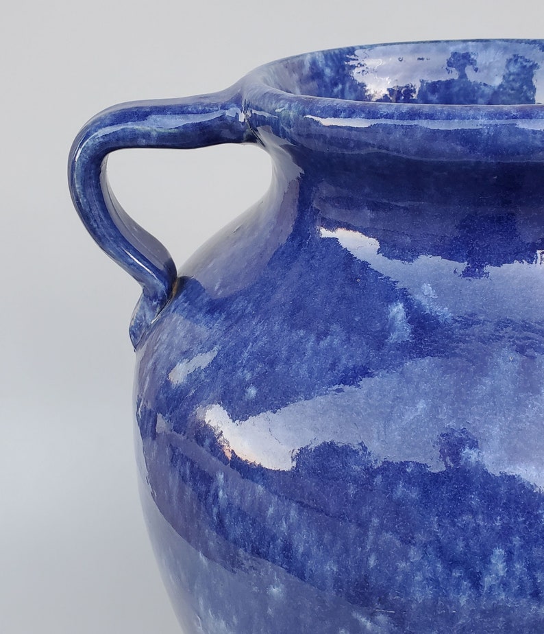 Op de afbeelding: Een blauwe keramische vaas met twee handvatten. De vaas heeft een gevlekt blauw glazuur met een licht iriserende afwerking.