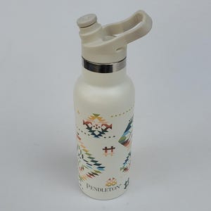May include: Off-white insulated water bottle with a flip-top lid and a silver band. The bottle features a colorful geometric pattern and the brand name "Pendleton" printed on the side. The bottle is designed to keep beverages cold.