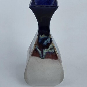 May include: A ceramic vase with a unique shape and color scheme. The vase has a square opening, a long, slender neck, and a wider, rounded base. The top is dark blue, transitioning to a black neck, with a white and brown base. The vase is approximately 8 inches tall.