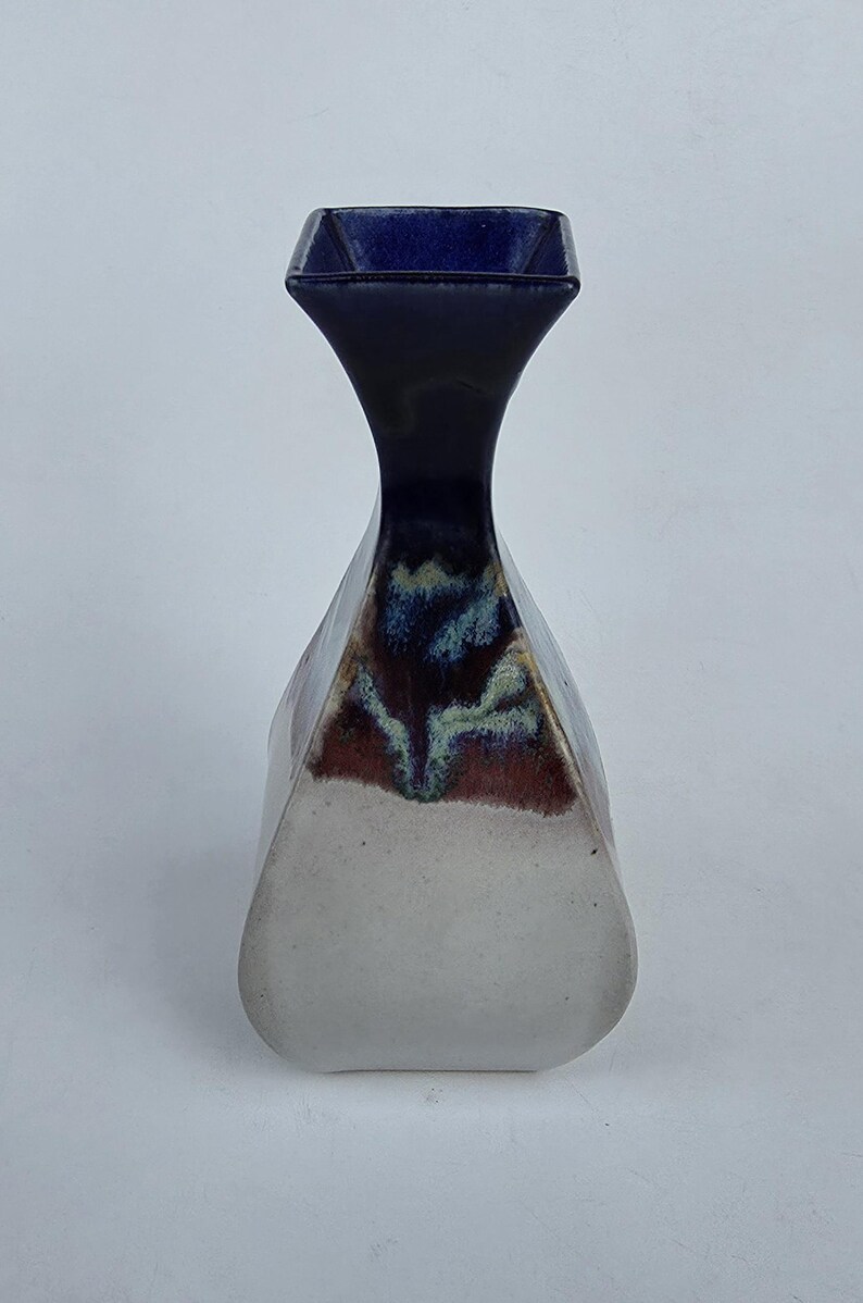 May include: A ceramic vase with a unique shape and color scheme. The vase has a square opening, a long, slender neck, and a wider, rounded base. The top is dark blue, transitioning to a black neck, with a white and brown base. The vase is approximately 8 inches tall.