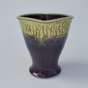 May include: A ceramic vase with a wide, flared opening and a narrow base. The vase is glazed in a deep purple color with a light green glaze dripping down the sides.