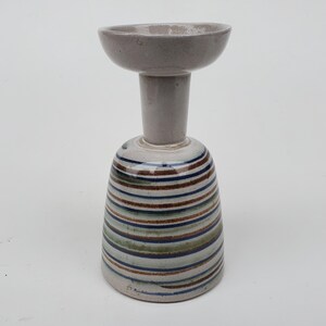 May include: A ceramic candle holder with a wide, shallow top and a striped base. The base features horizontal stripes in shades of brown, blue, and green. The top and stem are a solid, light gray color. The candle holder is a unique decorative item.