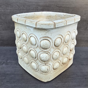 May include: A square ceramic planter with a glossy off-white glaze and a textured surface featuring a repeating pattern of raised circular medallions. The rim has a segmented border design, and the finish shows subtle brown speckling throughout.