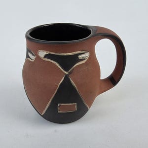 May include: A handmade ceramic mug with a brown exterior and a black interior. The mug features a geometric design in black and white, with a rounded handle. The mug is designed for hot beverages.