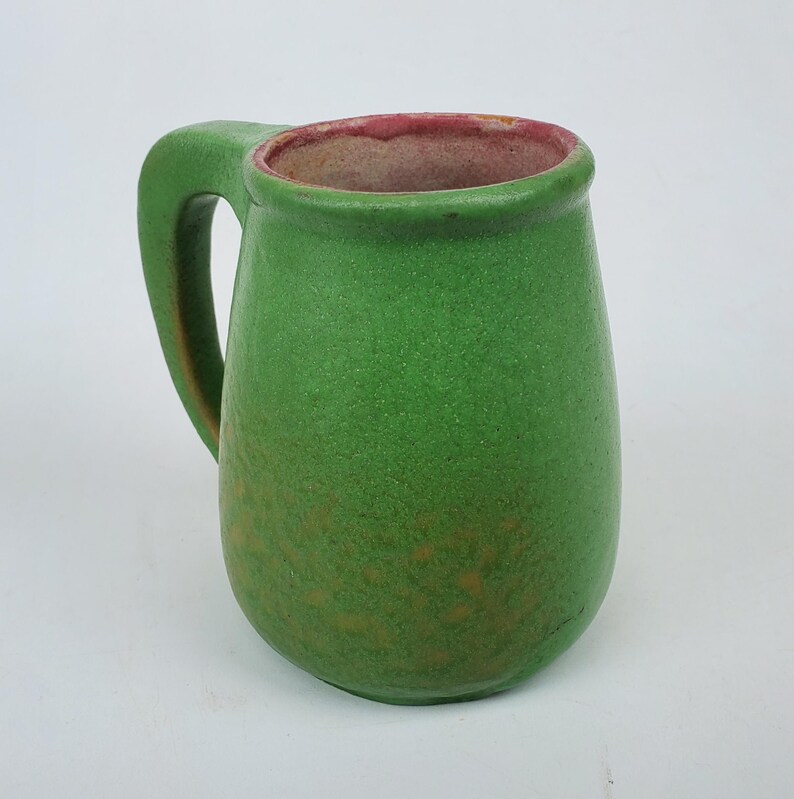 May include: A green ceramic mug with a large handle. The mug has a textured surface and a reddish-pink rim. The interior of the mug is a light brown color. The mug is a simple, yet elegant design.