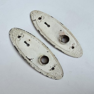 May include: Two oval-shaped metal door plates finished in distressed white paint. Each plate features a central circular opening for a knob, a keyhole above, and decorative embossed detailing along the edges with visible signs of wear.