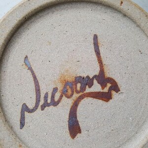 May include: Close-up of a ceramic plate with a brown signature that reads "Jucon" on the bottom.