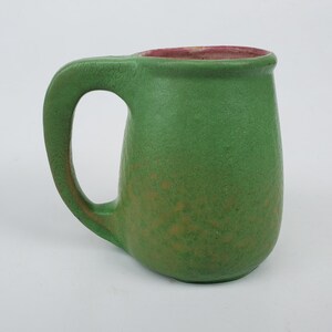 May include: A handmade ceramic mug with a green exterior and a reddish-pink interior. The mug has a rounded body, a large handle, and a textured surface. The mug is likely used for drinking beverages.