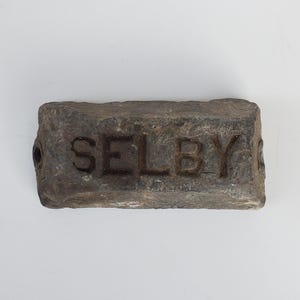 May include: A rectangular brick with the word "SELBY" embossed on its surface. The brick is a weathered grey colour, with a rough, textured finish. It features two small holes on the sides, and is likely made of clay.