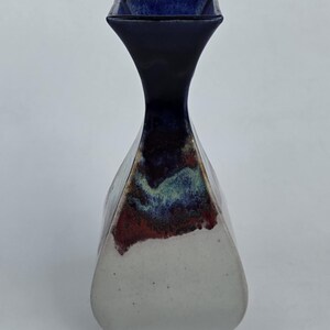 May include: A ceramic vase with a unique shape and color scheme. The top is a dark blue square, transitioning to a narrow neck. The body is white with a blend of red, blue, and green hues. A decorative piece for the home.