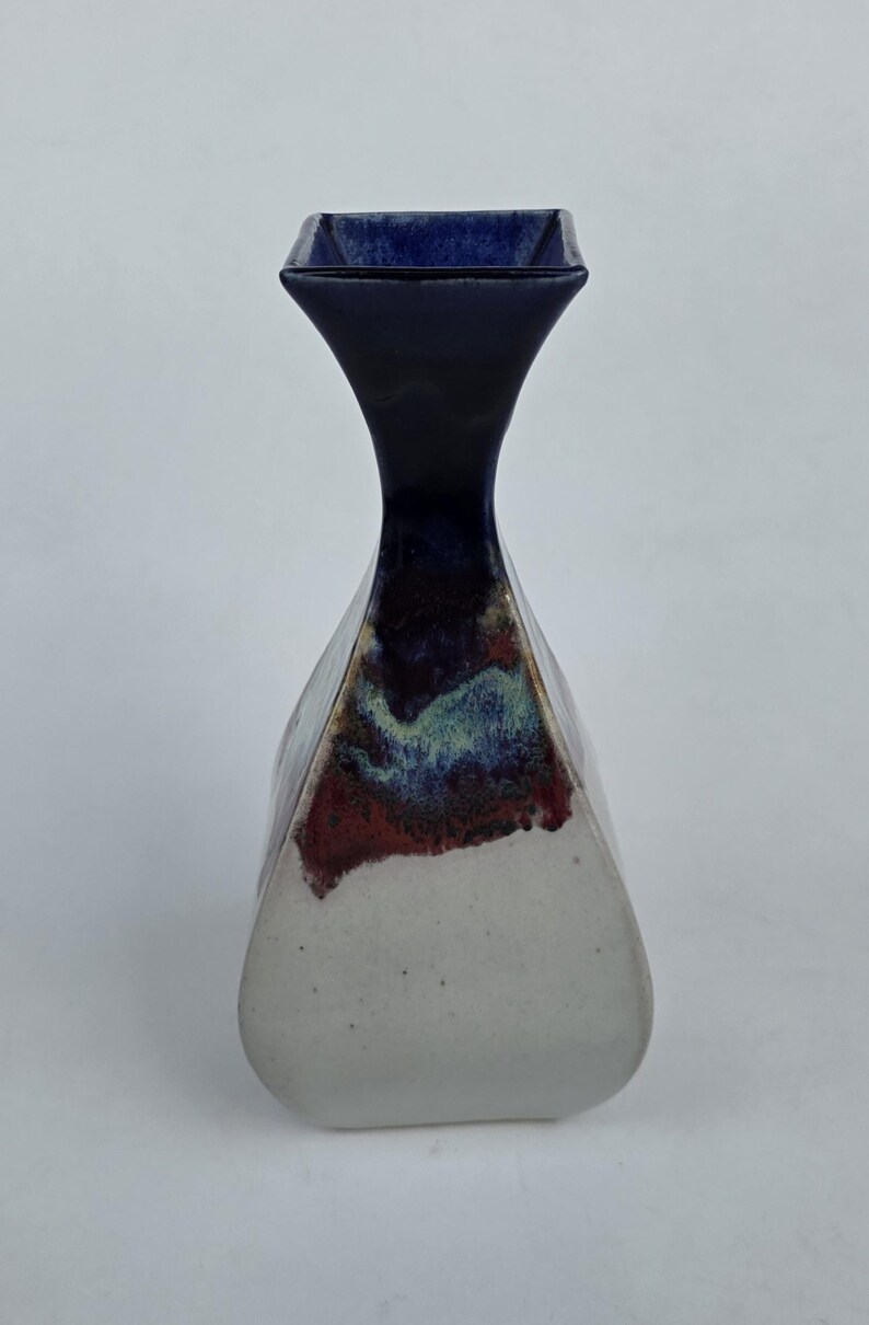 May include: A ceramic vase with a unique shape and color scheme. The top is a dark blue square, transitioning to a narrow neck. The body is white with a blend of red, blue, and green hues. A decorative piece for the home.