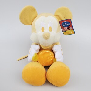 Puede incluir: Un juguete de peluche amarillo de Mickey Mouse sosteniendo un caramelo amarillo. El juguete tiene una cara blanca con ojos negros y una nariz negra. El juguete lleva una camisa blanca con rayas amarillas. El juguete tiene una etiqueta que dice "Disney Mickey Mouse Clubhouse Flavor of the Month!"