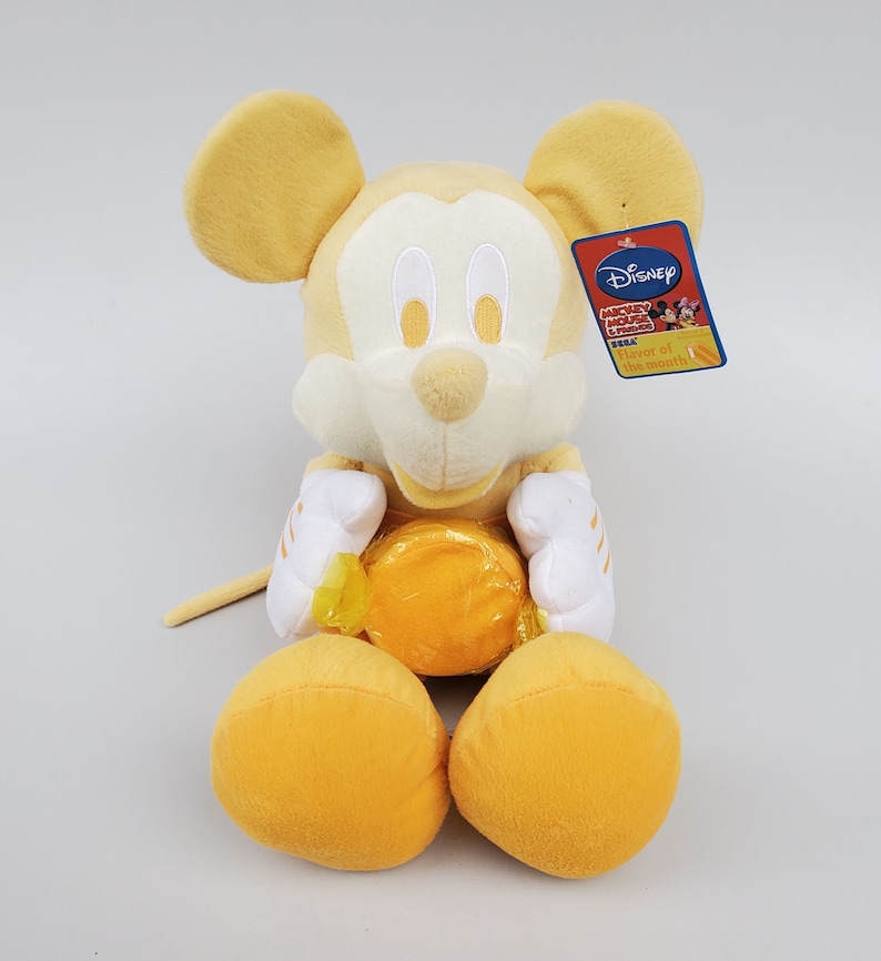 Puede incluir: Un juguete de peluche amarillo de Mickey Mouse sosteniendo un caramelo amarillo. El juguete tiene una cara blanca con ojos negros y una nariz negra. El juguete lleva una camisa blanca con rayas amarillas. El juguete tiene una etiqueta que dice "Disney Mickey Mouse Clubhouse Flavor of the Month!"