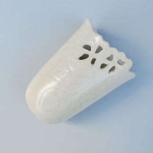 May include: White ceramic vase with a decorative cutout design. The vase has a textured surface and a slightly curved shape.