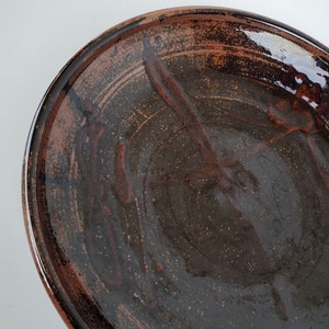 May include: A large, brown ceramic plate with a speckled glaze. The plate has a slightly raised rim and a smooth, glossy finish.