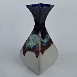 May include: A ceramic vase with a square top and a diamond-shaped body. The top is dark blue, transitioning to a multi-colored band of green, red, and purple, and then to a white base. The vase is a decorative home accent.