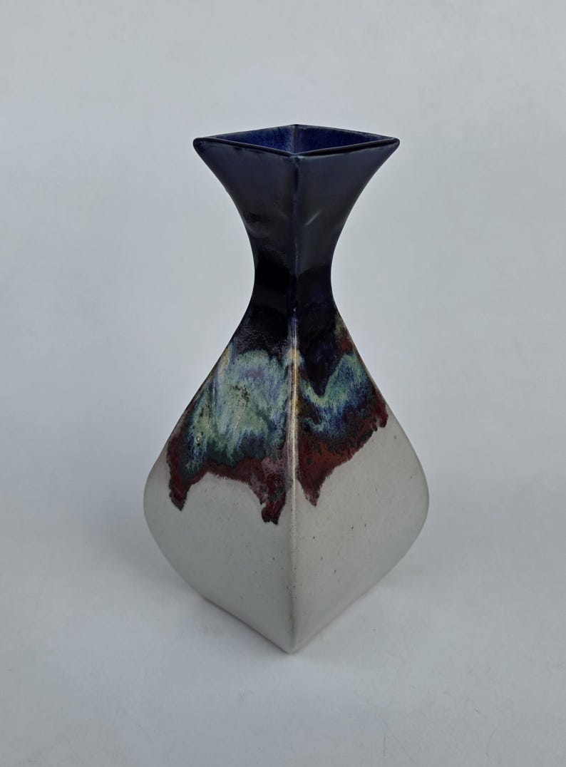 May include: A ceramic vase with a square top and a diamond-shaped body. The top is dark blue, transitioning to a multi-colored band of green, red, and purple, and then to a white base. The vase is a decorative home accent.