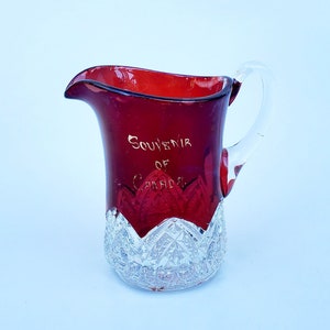 May include: A red glass pitcher with a clear glass handle. The pitcher has a clear glass base with a cut glass design. The pitcher is inscribed with the words "Souvenir of Canada".