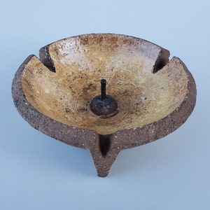 May include: A brown ceramic incense burner with three legs and a small hole in the center for incense sticks. The burner has a light brown glaze and a rough texture.