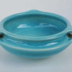 May include: A turquoise ceramic bowl with a glossy finish. The bowl has a rounded shape with two small handles on either side. The interior reflects light, and the overall design is simple and elegant.