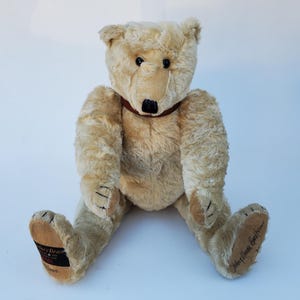 May include: A vintage-style teddy bear with light brown fur, black eyes, and a dark brown nose and collar. The bear is seated with its legs spread, and the paws have stitched details. The bear's foot has a label with text.