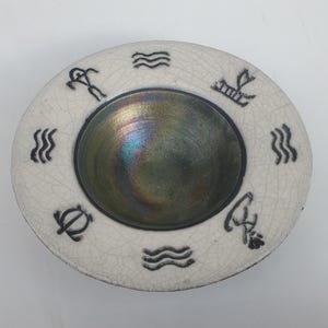 May include: A round, off-white ceramic dish with a central iridescent bowl. The rim features black symbols and wave patterns. The dish has a cracked glaze effect, adding texture. The bowl's interior reflects a spectrum of colors.