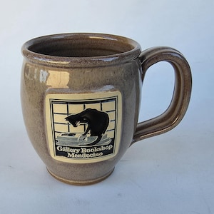 May include: A brown ceramic mug with a large handle. The mug features a rectangular label with a black cat illustration and the text "Gallery Bookshop Mendocino". The mug has a glossy finish.
