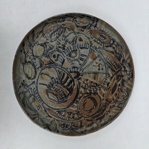 May include: A round ceramic plate featuring a textured surface with an abstract pattern of overlapping circles, curved lines, and geometric shapes in shades of brown, tan, gray, and black, with a dark brown rim encircling the entire piece.