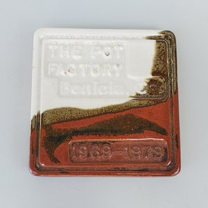 May include: A white ceramic tile with brown and red glaze. The tile has the text "THE POT FACTORY Bonita Ca." and "1969-1979" embossed on it.