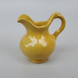 May include: A small, yellow ceramic jug with a rounded body and a curved handle. The jug has a glossy finish.