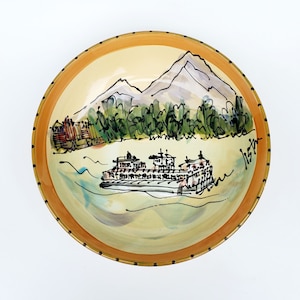 May include: A ceramic bowl with a hand-painted design of a ferry boat, mountains, and trees. The bowl has a yellow rim with black dots and a light yellow interior.