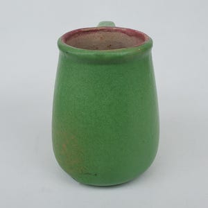 May include: A small, green ceramic pitcher with a handle. The pitcher has a textured surface and a rounded body. The interior rim is a reddish-brown color. The pitcher is likely handmade and suitable for serving beverages.