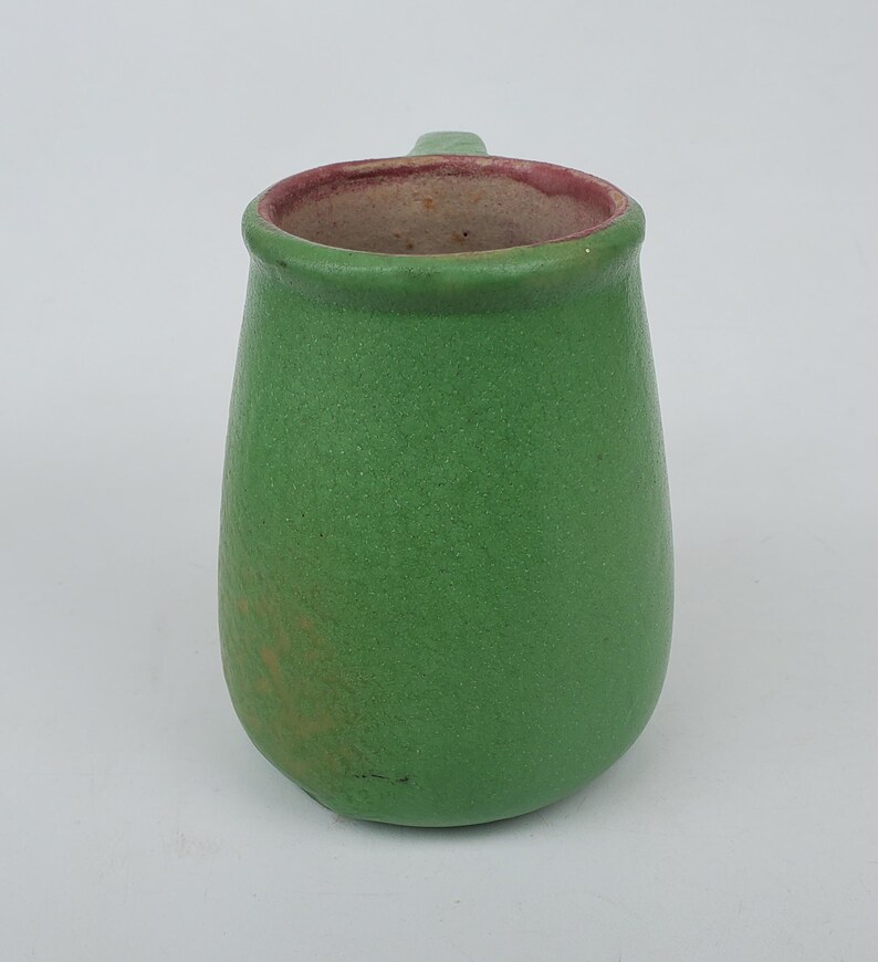 May include: A small, green ceramic pitcher with a handle. The pitcher has a textured surface and a rounded body. The interior rim is a reddish-brown color. The pitcher is likely handmade and suitable for serving beverages.