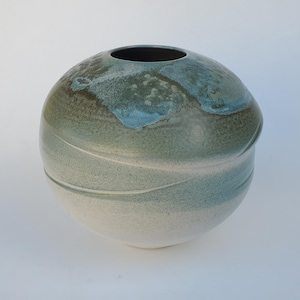 May include: A round ceramic vase with a light green and white glaze. The vase has a slightly uneven surface and a wide opening at the top.