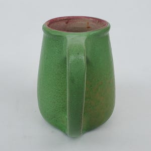 May include: A green ceramic pitcher with a handle. The pitcher has a textured surface and a red interior rim. The handle is a curved, solid piece. The pitcher is a solid green color with some variations in tone.