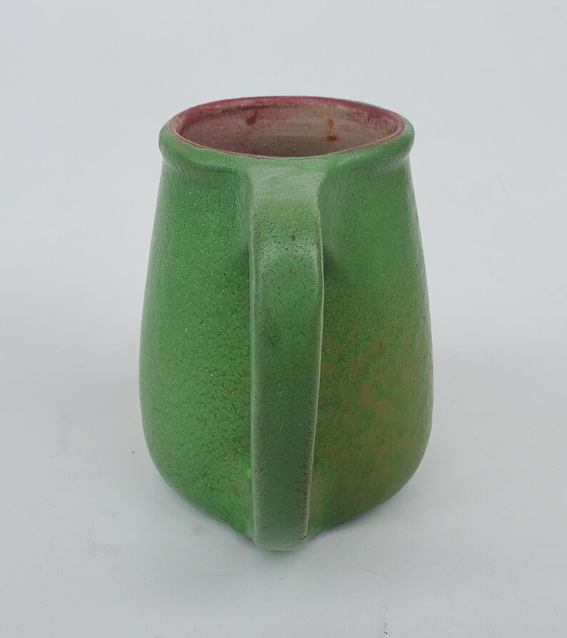 May include: A green ceramic pitcher with a handle. The pitcher has a textured surface and a red interior rim. The handle is a curved, solid piece. The pitcher is a solid green color with some variations in tone.