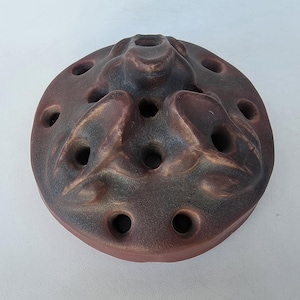 May include: A circular ceramic flower frog in brown and blue hues. The frog has a textured surface with multiple holes for arranging stems. The design incorporates raised, organic forms, creating a unique and functional piece for floral arrangements.
